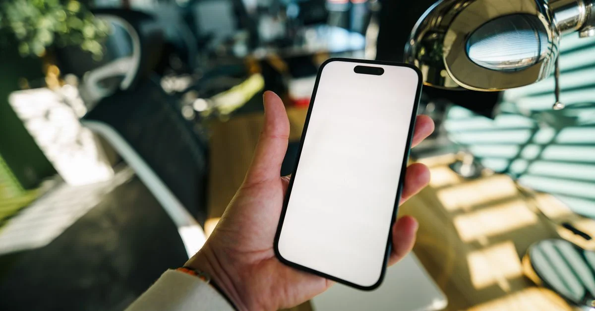 Une main tenant un smartphone avec un écran vide dans un espace de travail de bureau moderne et bien éclairé.