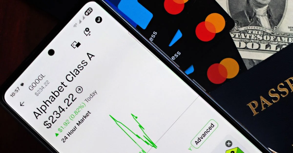 Un smartphone affichant le cours de l'action Alphabet à côté de cartes de crédit, d'argent liquide et d'un passeport.