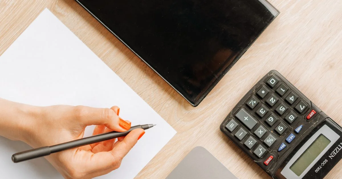 Vue de dessus d'un bureau minimaliste avec une main écrivant, une calculatrice et un appareil électronique.