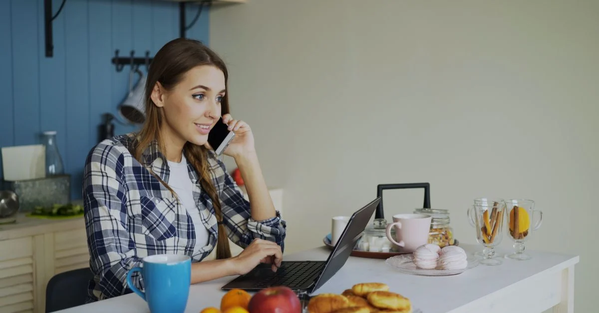 Jeune femme multitâche au téléphone et à l'ordinateur portable dans un cadre de cuisine confortable.