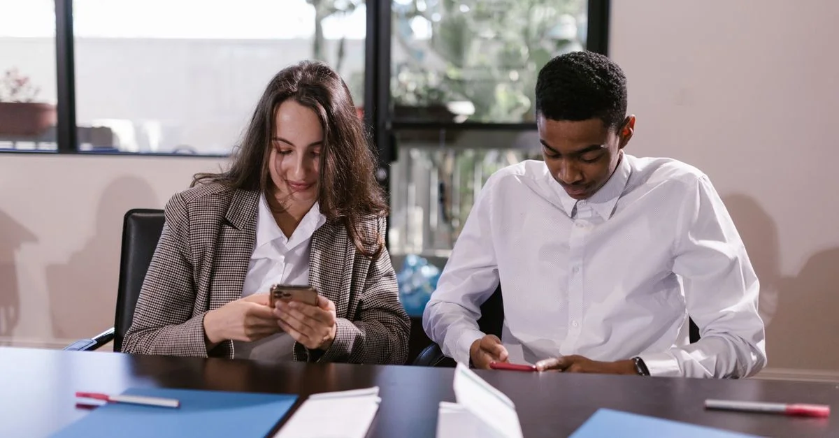 Deux jeunes professionnels concentrés sur leurs smartphones lors d'une réunion de bureau.