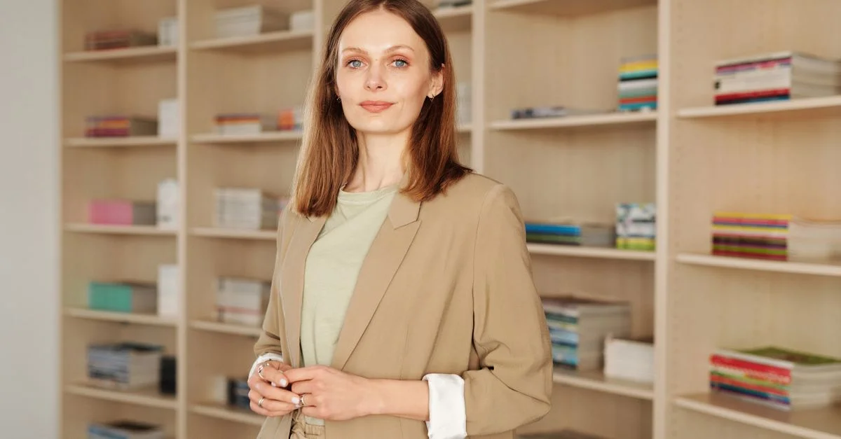 Femme élégante se tenant dans une bibliothèque, vêtue d'un blazer, entourée d'étagères de livres.